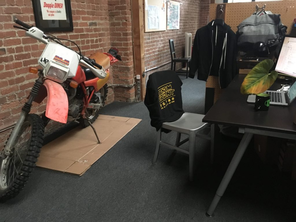 My desk at work, with a laptop and a mug of tea on top of it, and a large, old, somewhat ratty-looking dirt bike parked against the wall immediately behind it.