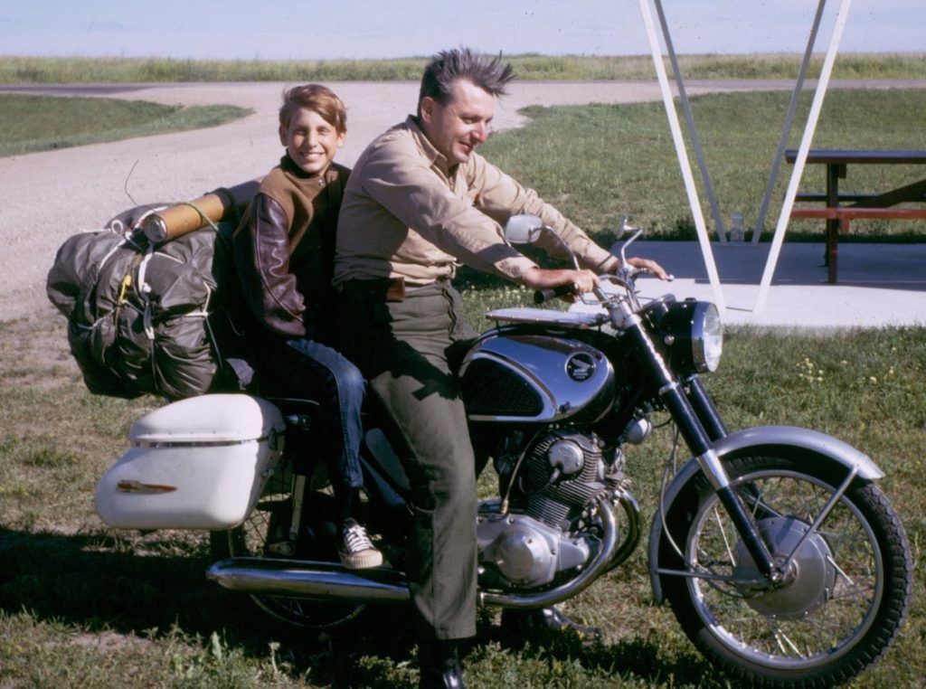 A man astride a parked motorcycle loaded with luggage, with a young boy riding pillion and smiling at the camera